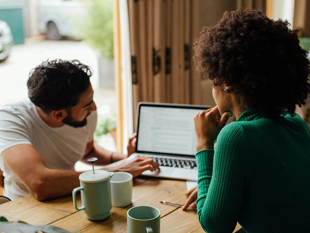 Zwei Personen sitzen mit Kaffeetassen an einem Holztisch und schauen gemeinsam in einem gemütlichen, sonnendurchfluteten Raum auf einen Laptop-Bildschirm. Zwei Personen sitzen mit Kaffeetassen an einem Holztisch und schauen gemeinsam in einem gemütlichen, sonnendurchfluteten Raum auf einen Laptop-Bildschirm.